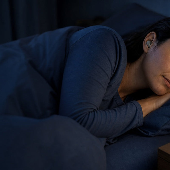 Side sleeper wearing a low-profile sleep earbud beside an open charging case in a dark bedroom